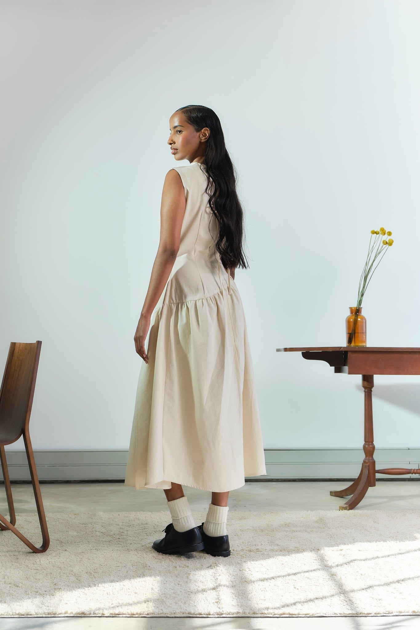 Woman in a beige dress standing in a room with a table and chair.