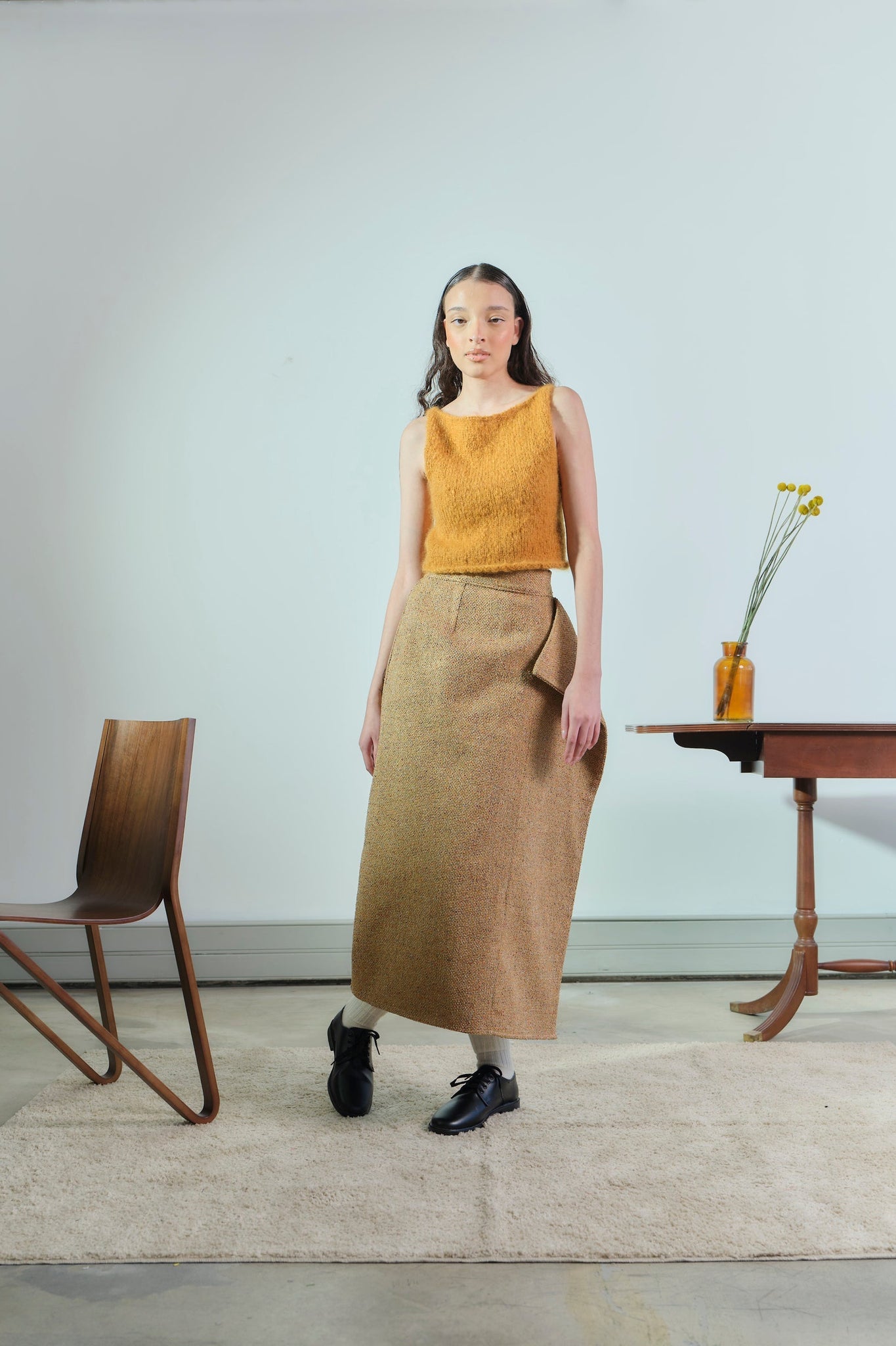 Woman in mustard top and brown skirt standing in a minimalistic room with a chair and table.