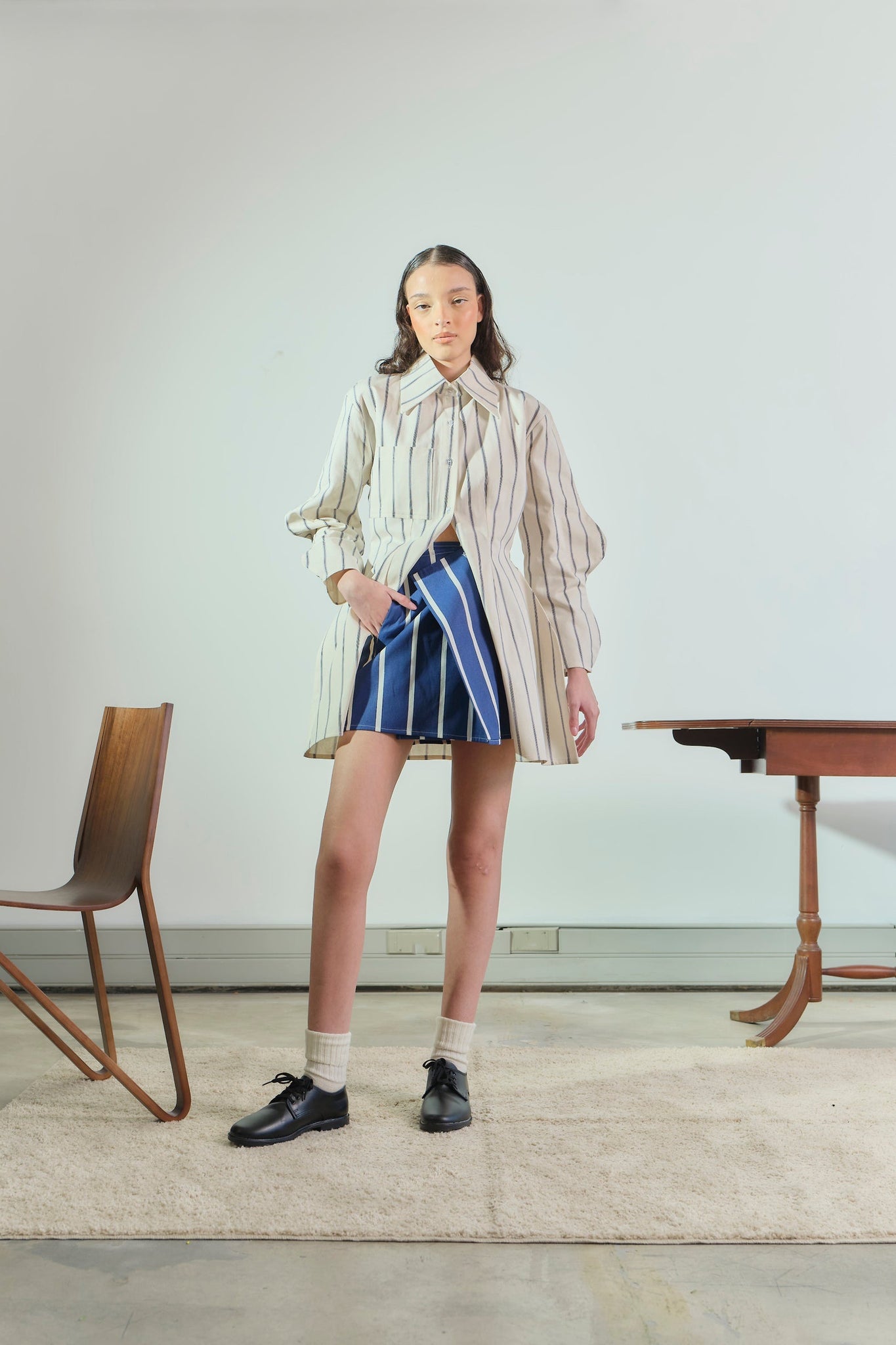 Woman in a striped shirt and blue skirt standing in a room with a chair and table.