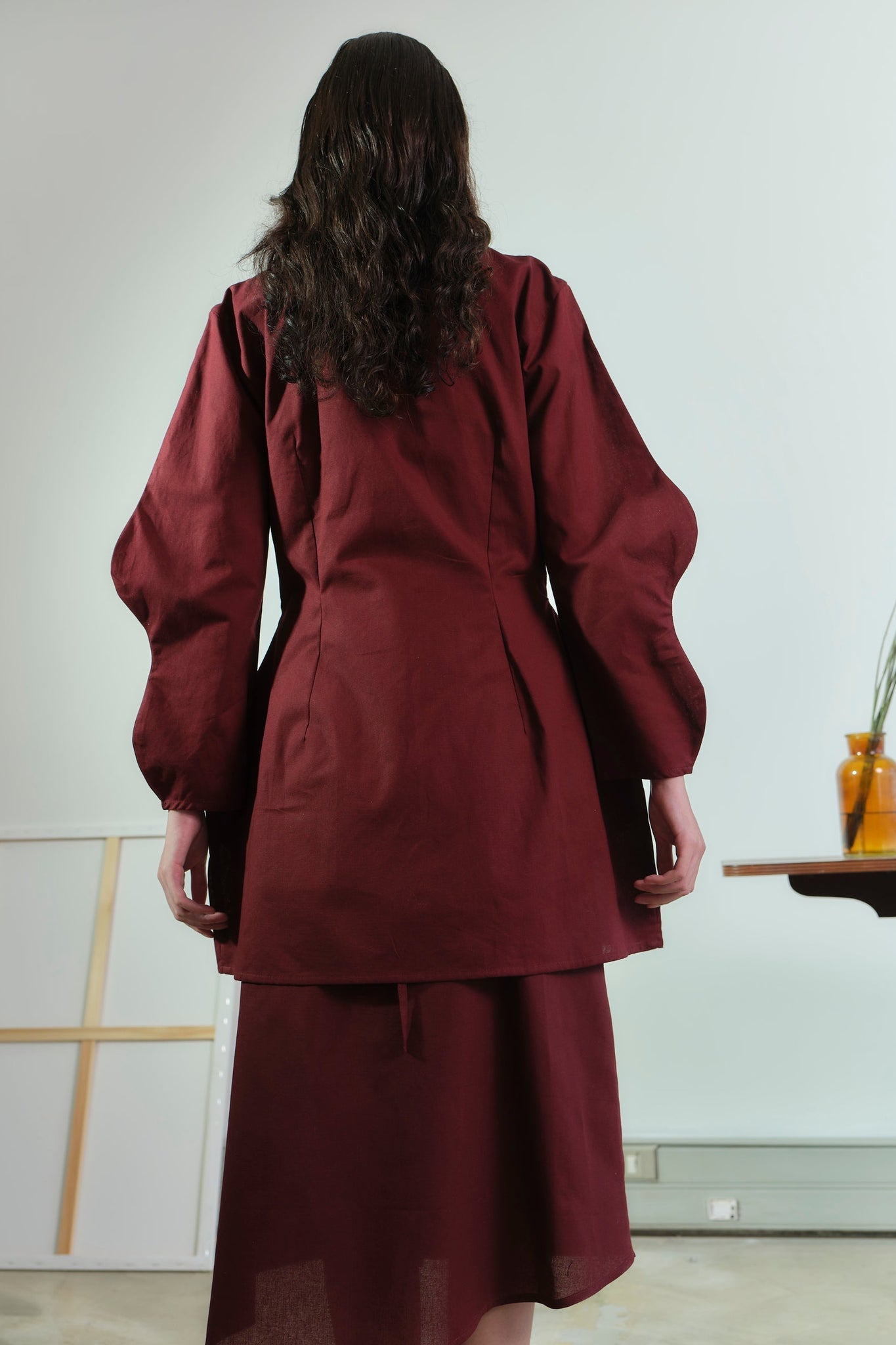 Person wearing a burgundy outfit standing in a minimalistic room with a white wall and wooden table.