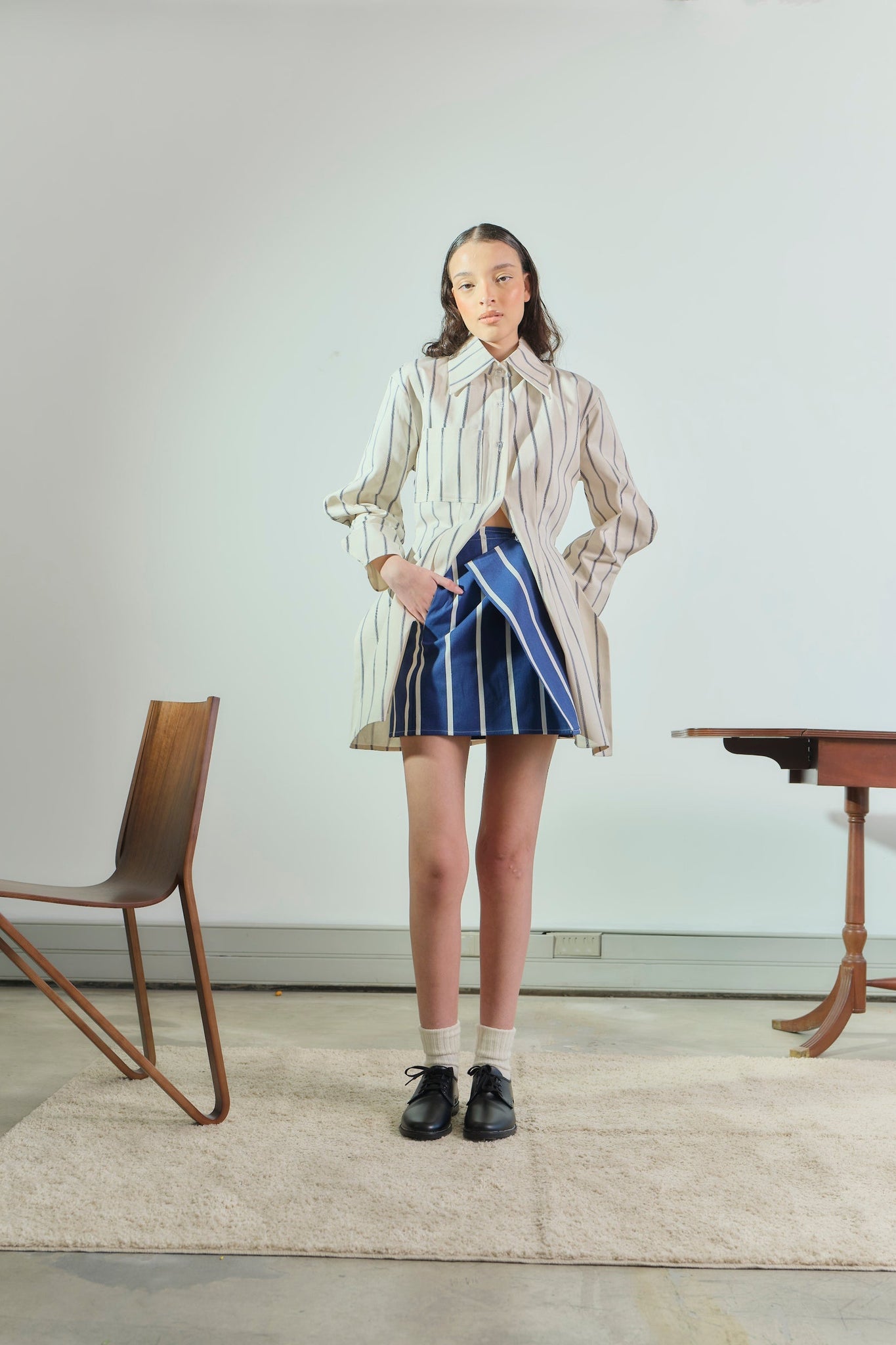 Person wearing a striped shirt and blue skirt standing in a minimalistic room with a chair and table.