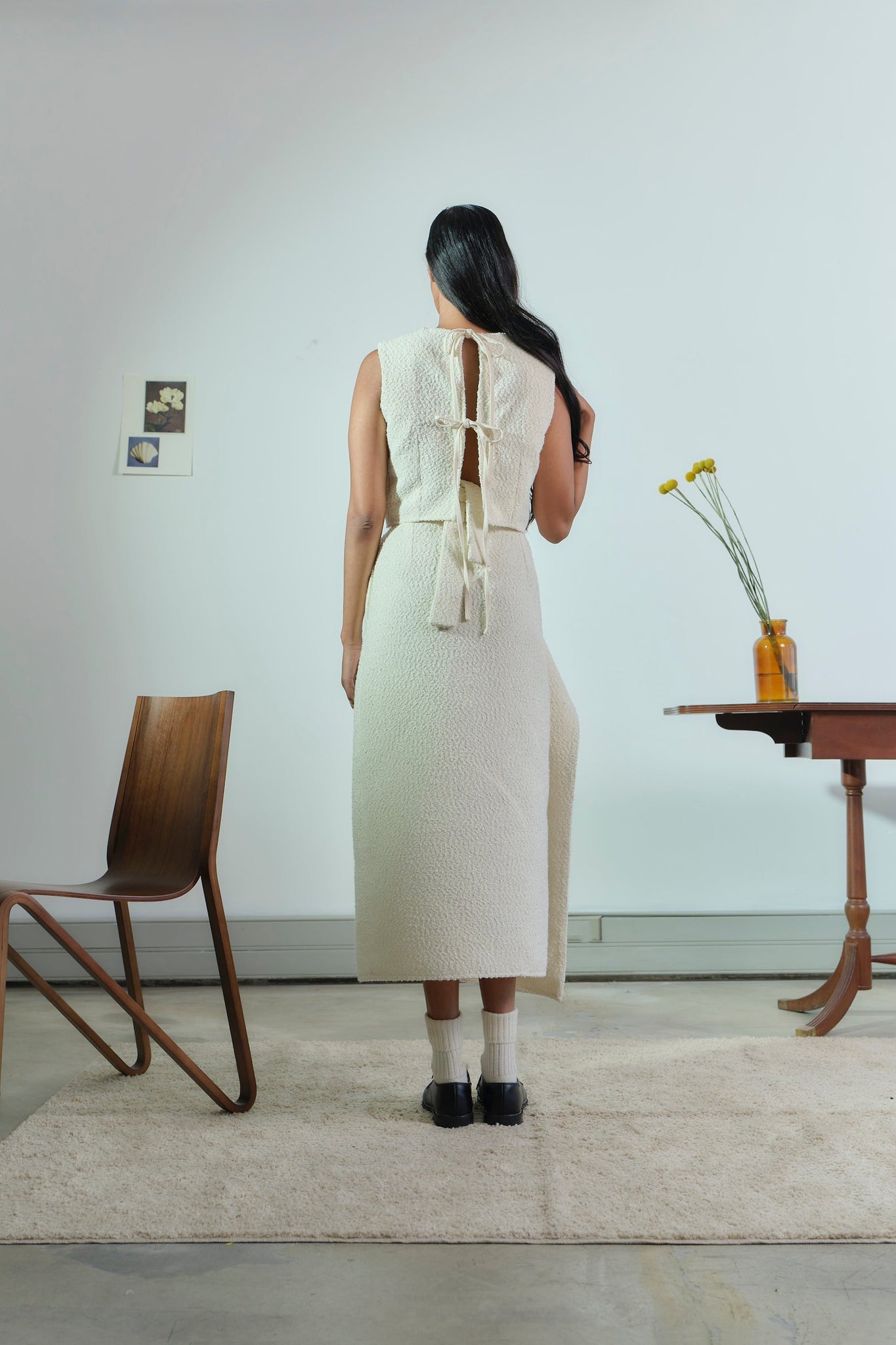 Woman in a white dress standing in a minimalistic room with a wooden chair and table.