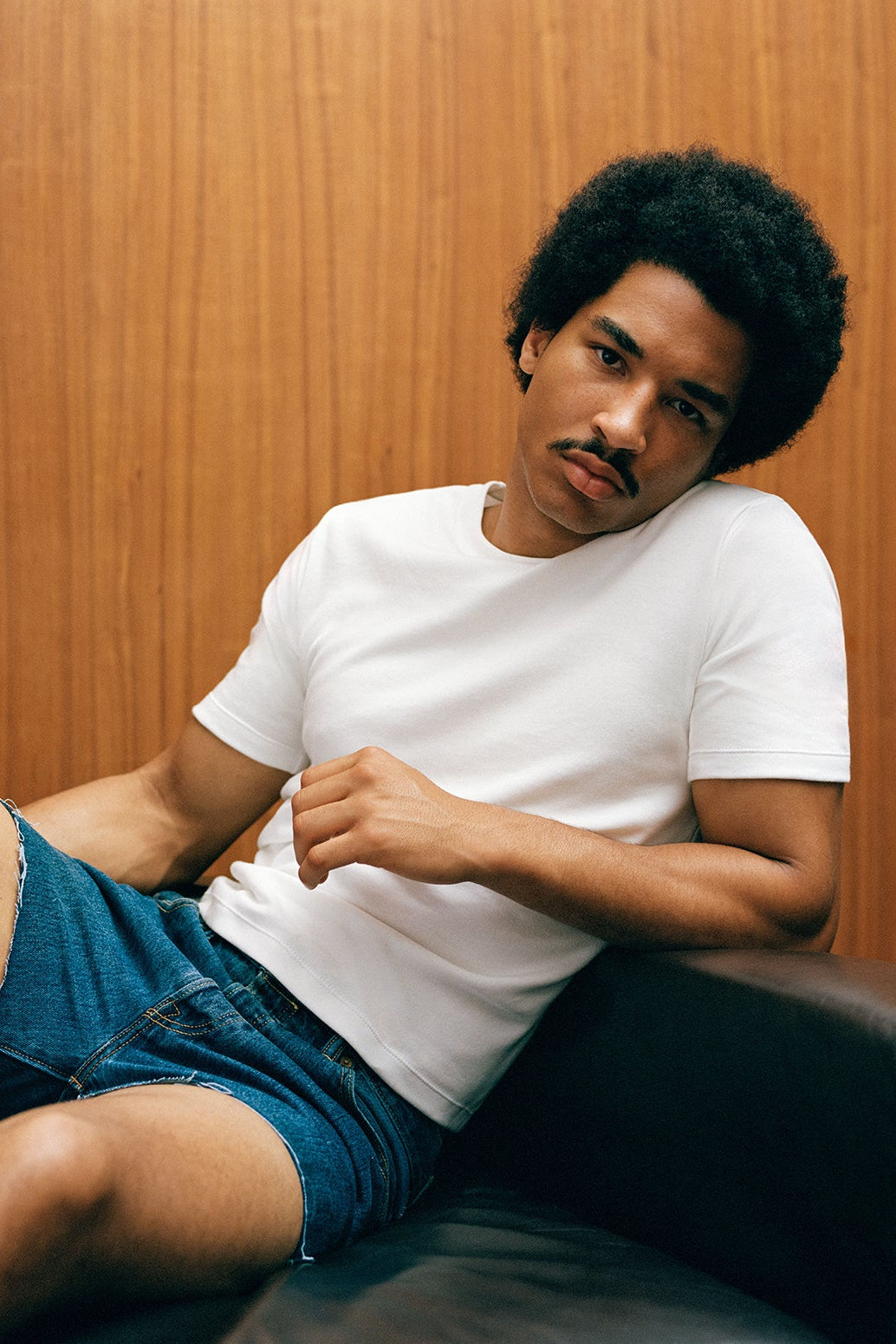 Man sitting on a couch wearing a white t-shirt and blue shorts against a wooden wall.