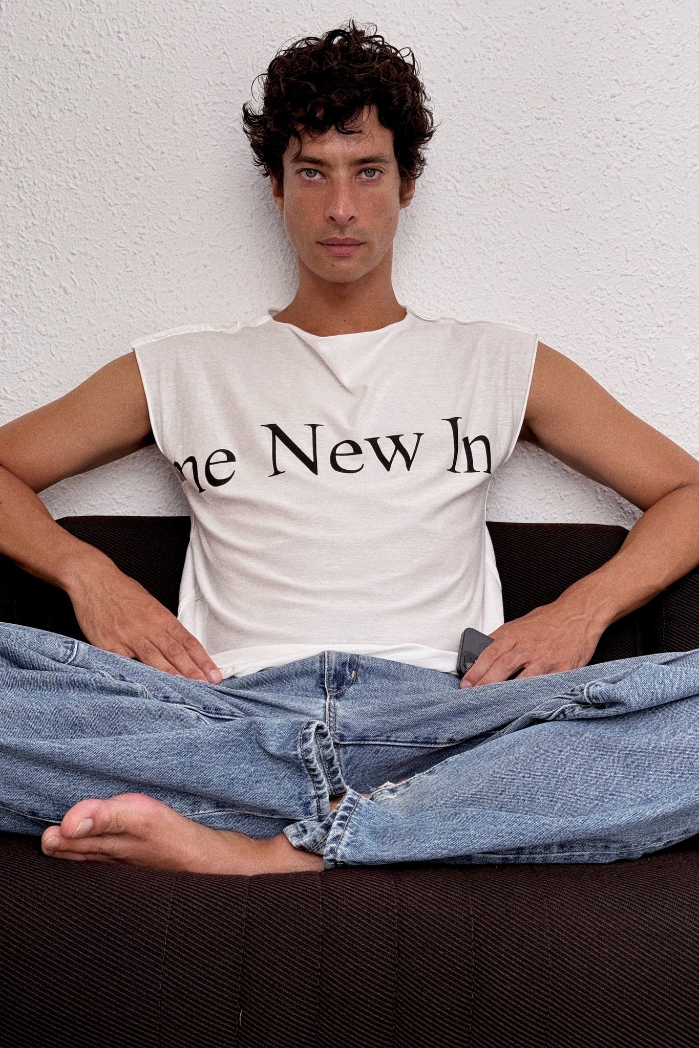 Man wearing a white sleeveless shirt with text and blue jeans sitting on a couch.