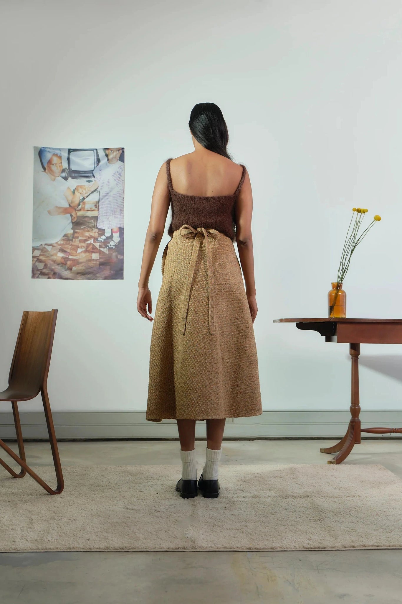 Woman wearing a brown dress standing in a room with a chair and table.